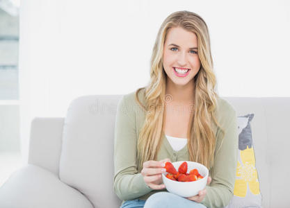 微笑的金发美女坐在舒适的沙发上吃草莓