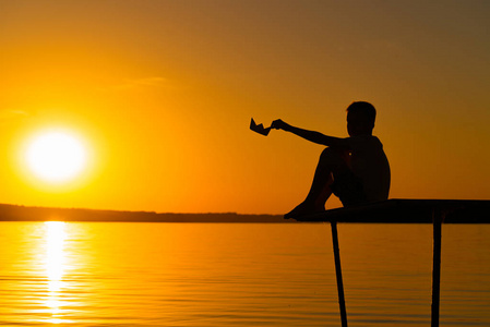 黄色纸船一个小男孩坐在桥上, 手里拿着纸船在河边日落.