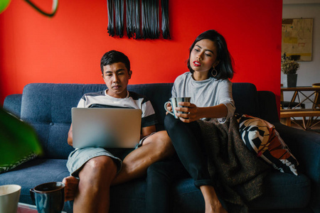 一个年轻的马来夫妇周末坐在家里。该名男子正在他的笔记本电脑上工作, 他的妻子正在检查她的智能手机