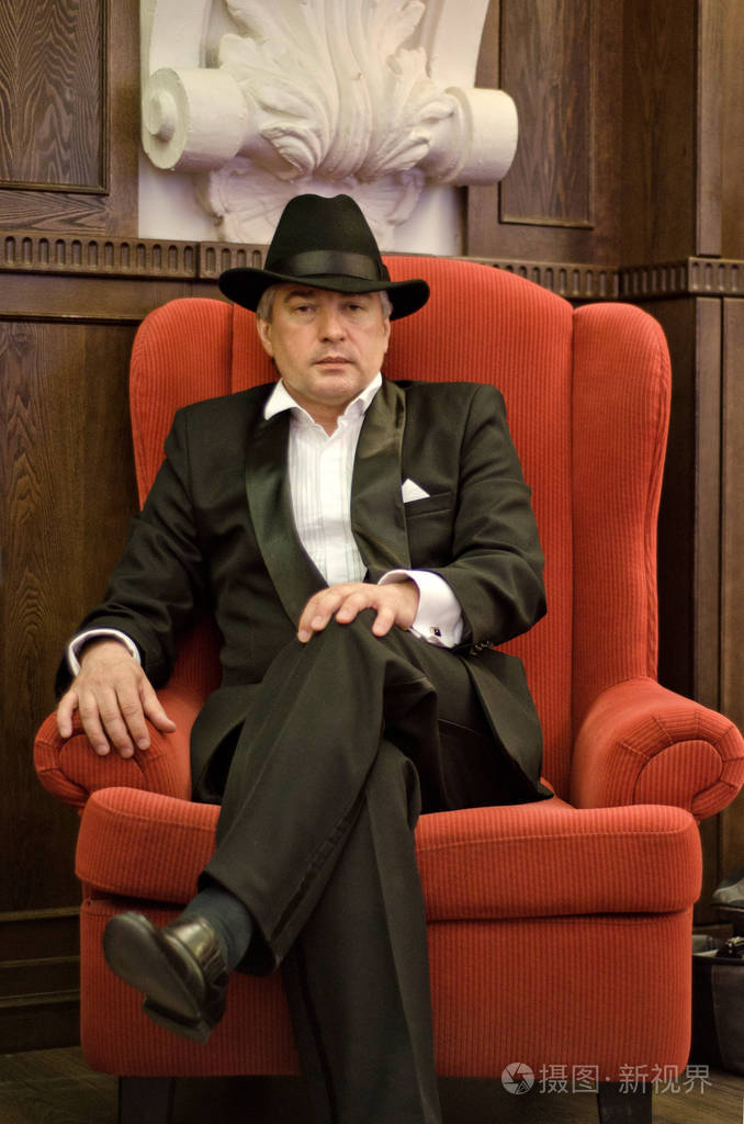 一个戴着黑色西装和帽子的灰色头发的老流氓坐在旧内饰的红色扶手椅上