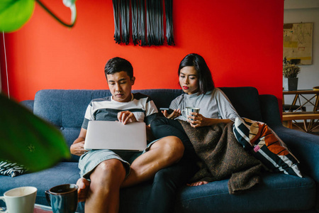 一个年轻的马来夫妇周末坐在家里。该名男子正在他的笔记本电脑上工作, 他的妻子正在检查她的智能手机