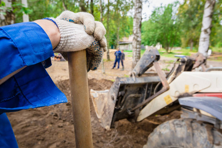 工人在蓝色 boilersuit 和工作手套与铲是在建筑工地上和模糊的现代挖掘机, 在施工现场进行开挖工程在重建地方