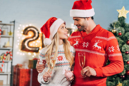 一对夫妇在圣诞老人的帽子与香槟眼镜持有存钱罐在圣诞节的储蓄