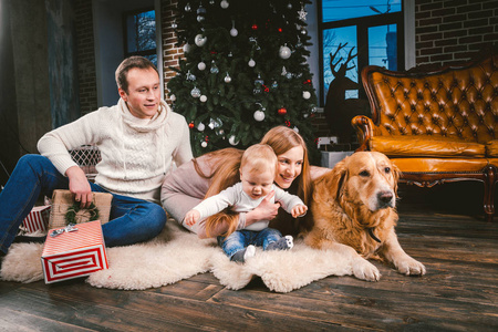 主题圣诞节和新年家庭圈子和家庭宠物。妈妈爸爸和孩子1岁的白种女人坐在圣诞树旁边的礼物和大狗品种拉布拉多金猎犬