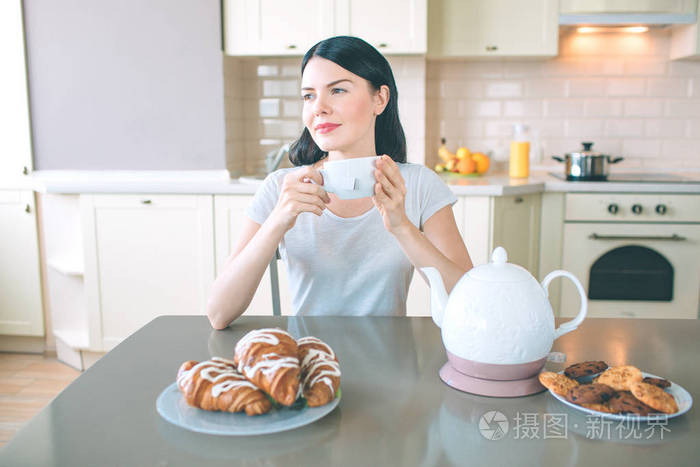 梦幻般的女人坐在餐桌前, 看起来很对。她手里拿着白杯子。桌上有 croissans 和饼干的水壶和盘子。