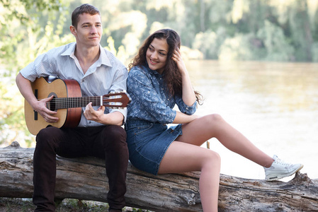 年轻夫妇坐在河边的原木上玩吉他, 夏天的自然, 灿烂的阳光, 阴影和绿叶, 浪漫的感觉