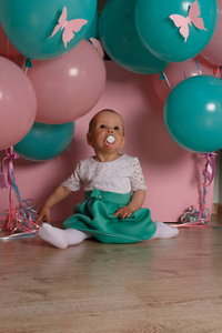 一个小女孩坐在地板上, 粉红色的背景, 靠近气球, 用奶嘴. 庆祝。生日, 转了一岁。穿白色和蓝色。小女孩与奶嘴
