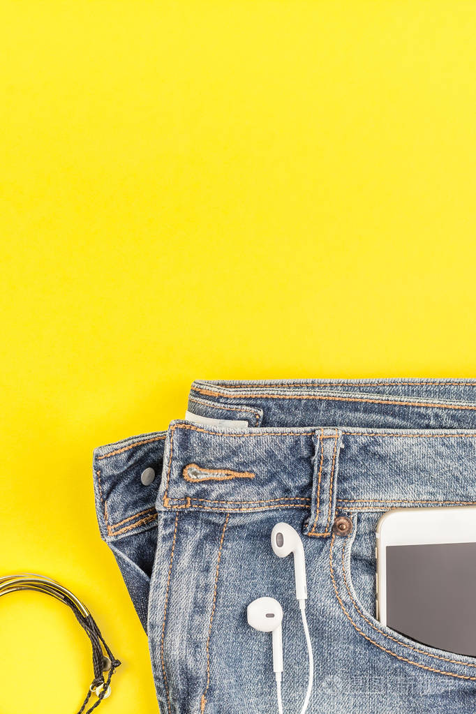 平躺在蓝色牛仔裤和智能手机与耳机在大胆的黄色纸背景与复制空间。女性休闲服装的头顶景色。时髦的时髦的看顶部视图