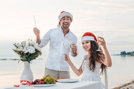 新婚夫妇在圣诞老人的帽子庆祝新年香槟杯和烟花在海滩上