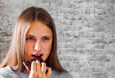 年轻的黑发女孩的肖像在灰色墙壁背景下吃巧克力