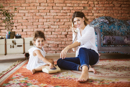 年轻的微笑的母亲和小可爱的女儿愉快地看在照相机演奏与复古电话一起在地毯在家