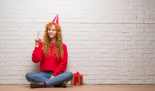 年轻的红头发妇女坐在砖墙上戴着生日帽惊讶的想法或问题指向手指与快乐的脸, 第一