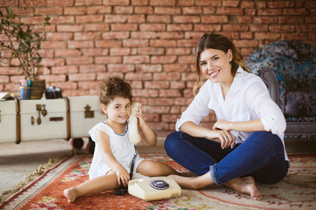 美丽的母亲与小女儿愉快地看在照相机玩复古电话在地毯在家