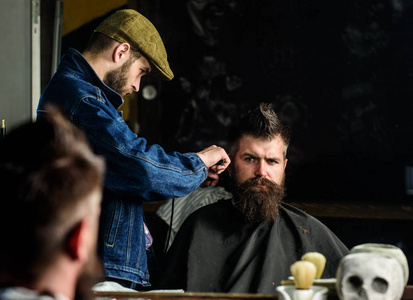 时髦的客户得到理发。理发师与理发剪工作在发型为人与胡子, 理发店背景。理发师发型的野蛮胡子客户端与剪刀。理发概念