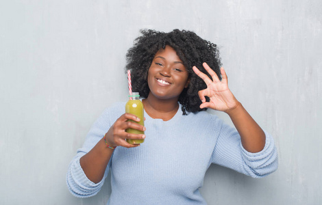 年轻的非洲裔美国妇女在灰色垃圾墙上喝排毒绿色果汁做 ok 标志与手指, 优秀的标志