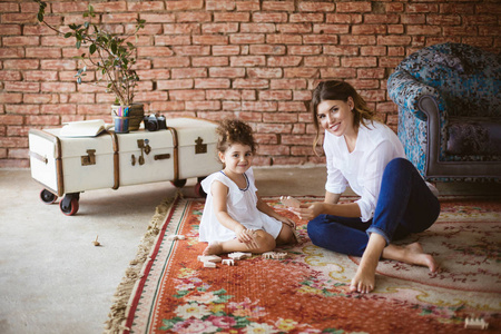 年轻漂亮的母亲在白色的衬衫和小可爱的女儿在白色的礼服高兴地看着相机玩木玩具一起在舒适的家地毯