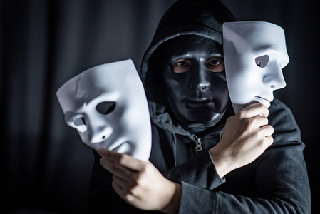 神秘的帽衫人戴着黑色面具, 手里拿著两个白面具。匿名社交屏蔽。主要抑郁症或躁郁症。万圣节概念
