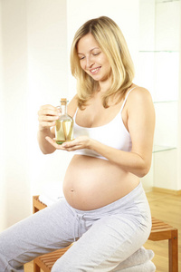 金发怀孕的妇女穿着白色内衣持有瓶与身体油按摩她的腹部在浴室