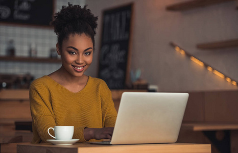 美丽的美国黑人女孩在休闲服装是使用笔记本电脑, 看着相机和微笑, 而坐在咖啡馆