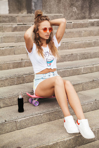 美丽的年轻女子在太阳镜下喝着五颜六色的鸡尾酒从瓶子, 坐在楼梯上的街道上。时尚的发型。生活方式肖像