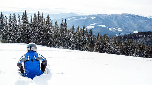 年轻英俊的男子在冬季运动服看着, 戴着大镜子滑雪面具. 穿着蓝色滑雪服的男子坐在山顶上的雪上。