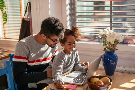 非洲裔美国父亲和女儿看室内便携式计算机上的东西。她正坐在他的大腿上。他们是微笑和快乐的感觉