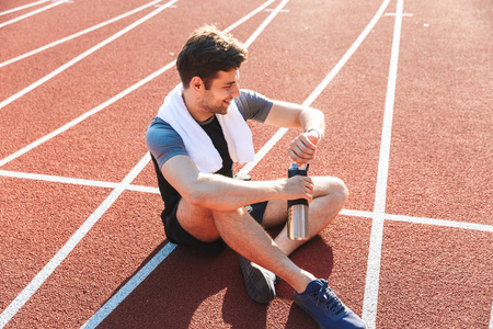 疲惫的运动员在体育场跑完, 休息, 拿着水壶, 看着 smartwatch