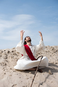 快活的耶稣在长袍和红色窗扇坐在莲花位置与举起的手和谈话与上帝在沙子在沙漠