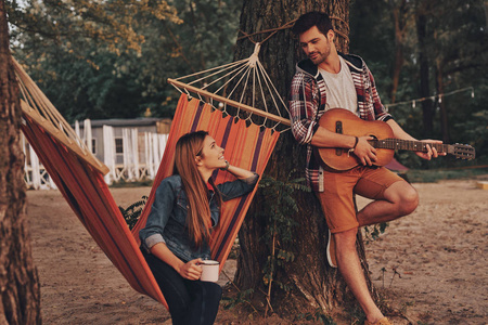 英俊的年轻人弹吉他为美丽的女朋友在户外在森林里, 妇女坐在吊床与杯子