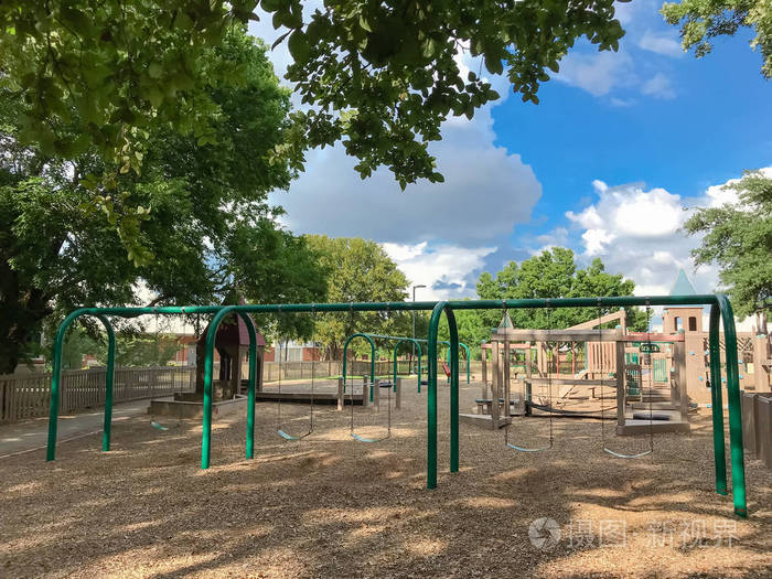 在美国德克萨斯州科佩尔的大型树木繁茂的公共木制儿童游乐场上设置拱秋千。游戏集结构环绕着夏日的叶子绿云蓝天