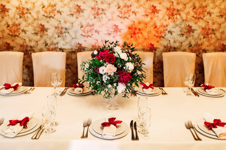一张漂亮的桌子, 墙上有一束别致的花束。白色餐巾与红色弓在板材和餐具用玻璃