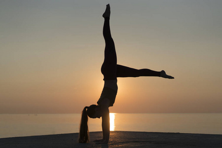 侧面看到一个女人的剪影, 站在她的手。健康灵活的女孩在日出时做瑜伽。在西班牙的夏日假期, 在宁静的海滩上练习倒立姿势的女孩