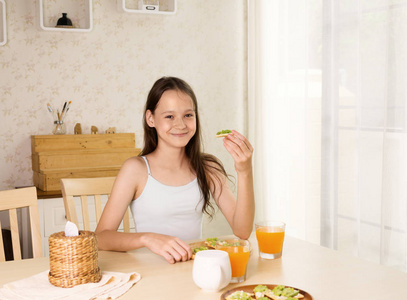 可爱的微笑青春期女孩有健康的早餐 鳄梨三明治和橙汁。健康的生活方式概念, 素食食品。保健品.客厅室内背景