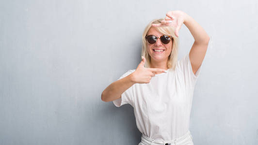 成年白人妇女在灰墙上戴着太阳镜, 微笑着用手和手指与快乐的脸做框架。创意与摄影理念