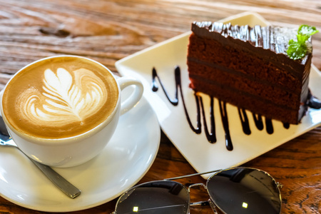 杯拿铁艺术咖啡匙与温暖的光亮美丽纹理背景装饰着太阳镜和一块巧克力软糖蛋糕的棕色树皮上板