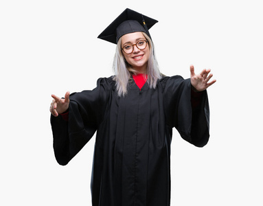 年轻的金发女子穿着毕业制服在孤立的背景看着相机张开双臂微笑拥抱。快乐的表达拥抱幸福