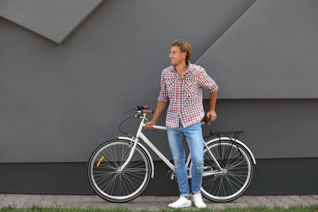 在室外灰色墙壁附近的英俊的年轻人与自行车