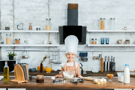 可爱的孩子在厨师帽子和围裙烹调面团和微笑在照相机