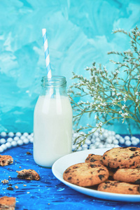 饼干巧克力与在蓝色背景花附近的有机牛奶瓶。健康早餐的概念。简约.春季食品