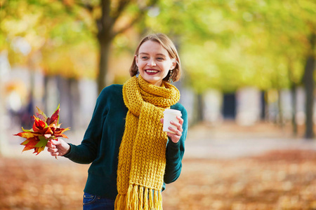 愉快的小女孩在黄色围巾与咖啡去漫步在秋天公园在一个明亮的秋天天