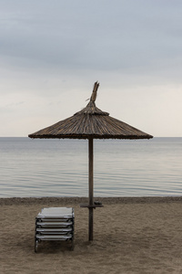 在沙滩上日光浴的芦苇阳伞。伞和日光浴浴床上阴沉的天气和平静的海洋。芦苇的伞和安乐椅上多云的天空背景