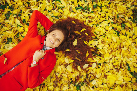 美丽快乐的女孩与长波浪头发装饰与黄叶铺设在秋季森林举行大衣衣领与手。愉快的美丽的女孩在红色外套在美丽的秋天天