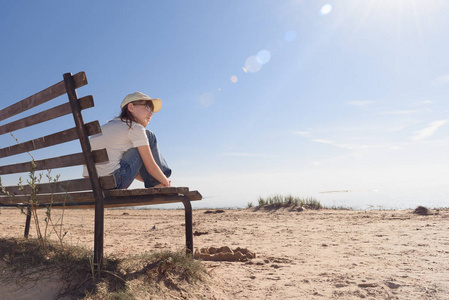孤独的女孩戴着眼镜, 坐在海边的长凳上, 悲伤地望向远方.照片