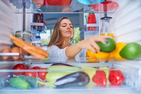 妇女站在打开的冰箱前, 并采取鳄梨。冰箱里装满了杂货