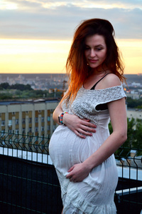 年轻美丽和快乐怀孕红发女孩的日落和市看观提出的未来的母亲和她怀孕在浪漫的白色连衣裙