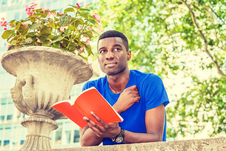 成功之路。年轻的非洲裔美国大学生在纽约学习, 穿着蓝色短袖 t恤, 站着, 向后弯曲在校园墙上, 手捧红书, 思考