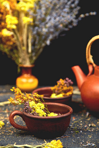 杯草药茶tutsan, 艾树, 牛至, 蜡菊, 薰衣草附近棕色茶壶在黑暗的木质背景。凉茶。干草药和花卉, 草药