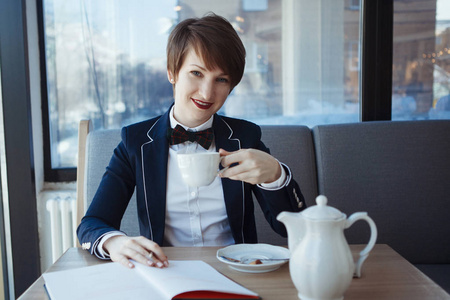 年轻的商业妇女在西装与弓领带是喝茶和阅读一本书在午休时间