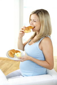 愉快的怀孕妇女坐在扶手椅和吃羊角面包从盘子在家里
