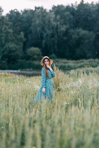 美丽的俄罗斯女孩在帽子和夏季礼服, 走在田野上的草地上的腰部。快乐的旅行者, 收集草药, 微笑。森林中的日落